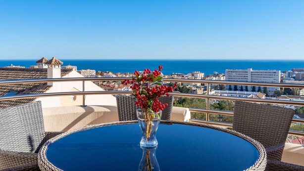 A balcony with a table and chairs, a vase with red berries, and a cityscape with the sea in the background.