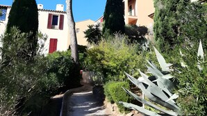 Un sendero atraviesa un jardín con diversos árboles y arbustos, rodeado de edificios con contraventanas rojas y un cielo azul despejado.
