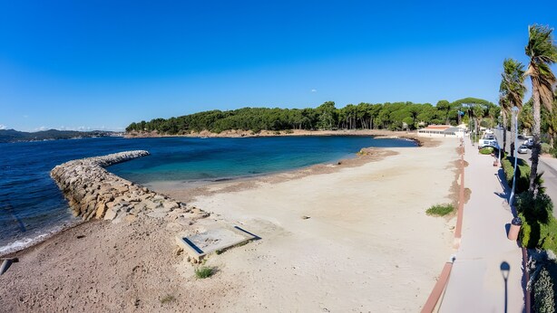 Una playa costera con un espigón de piedra, un paseo marítimo pavimentado y una fila de coches aparcados.