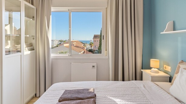 Quarto com cama, janela com vista para edifícios e o mar, guarda-roupa e mesa de cabeceira com abajur.