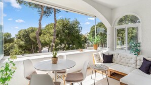 Une terrasse ensoleillée avec un canapé blanc, une table ronde et des chaises, offrant une vue sur un paysage verdoyant et luxuriant.
