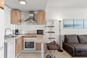 A modern kitchen with a stove, oven, and coffee maker, a dining area with a table and chairs, and a sofa in a living area with a cityscape view. - Apartment 'Gran Terraza con Vistas al Mar A111' with Wi-Fi and Air Conditioning (Mijas Costa)