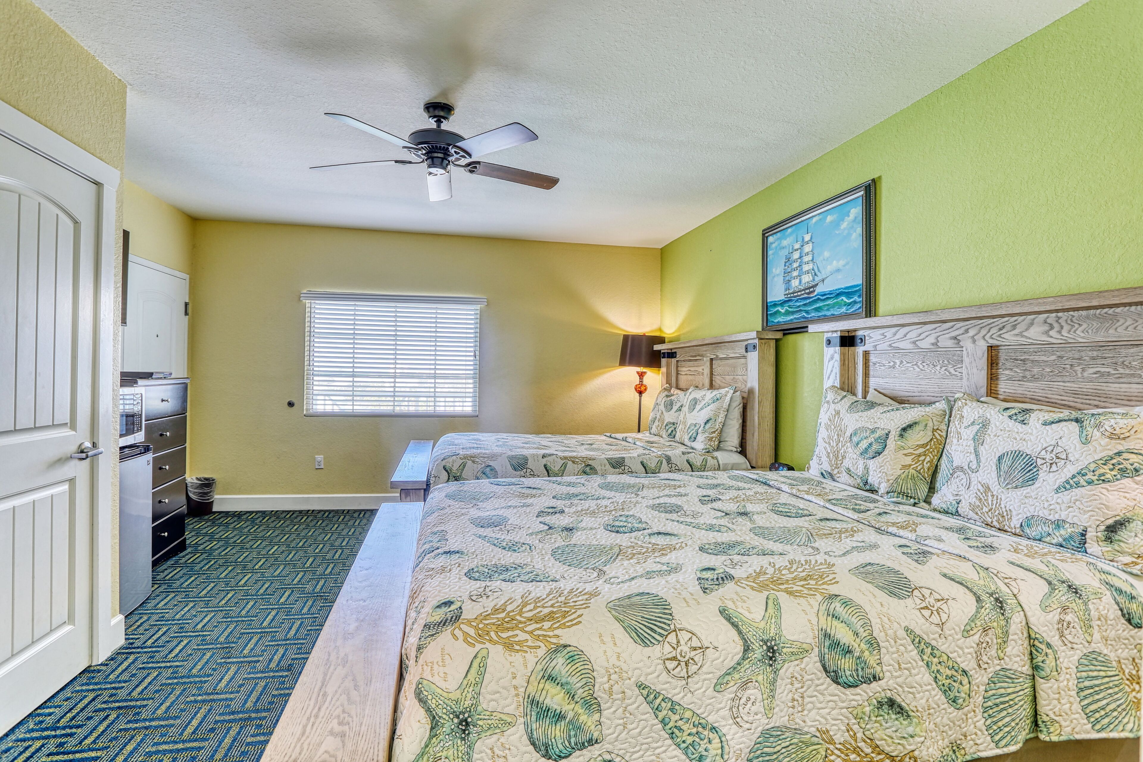 A bedroom with a bed, a ceiling fan, a window with blinds, and a nightstand with a lamp.