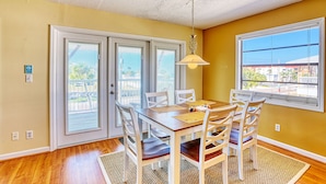 A dining area with a wooden table and white chairs, a hanging light fixture, and a view through a window.
