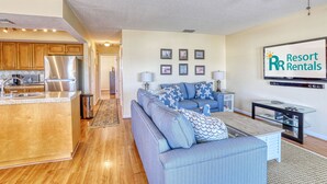 A modern living room with a sofa, coffee table, and a TV. There is a kitchen area with a refrigerator and cabinets.