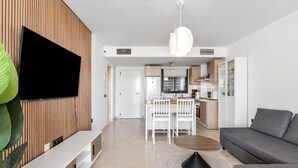 Una sala de estar moderna con un televisor de pantalla plana, un sofá gris, una mesa de centro y un comedor con sillas, con una cocina al fondo.