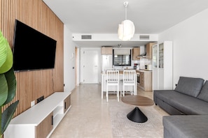 A modern living room with a flat-screen TV, a grey sofa, a coffee table, and a dining area with chairs and a kitchen in the background.