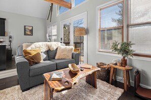 A modern living room with a grey sofa, wooden coffee table, and a large window with blinds.