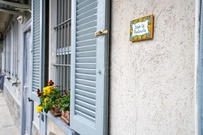 Une façade d’immeuble avec une fenêtre ornée de fleurs jaunes et rouges, une enseigne fixée au mur portant l’inscription « Casa la Fontanella », et une surface murale à texture.