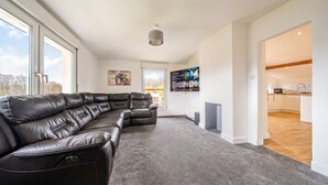 A modern living room with a sectional sofa, a flat-screen TV, and a view to the kitchen. - Family-friendly countryside home with hot tub, swing set and trampoline (Battle)