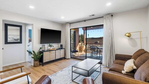 A modern living room with a flat-screen TV, a sofa, a coffee table, and a dining area visible through glass doors. - Avondale Hideaway - Condo for 6 (Branson)