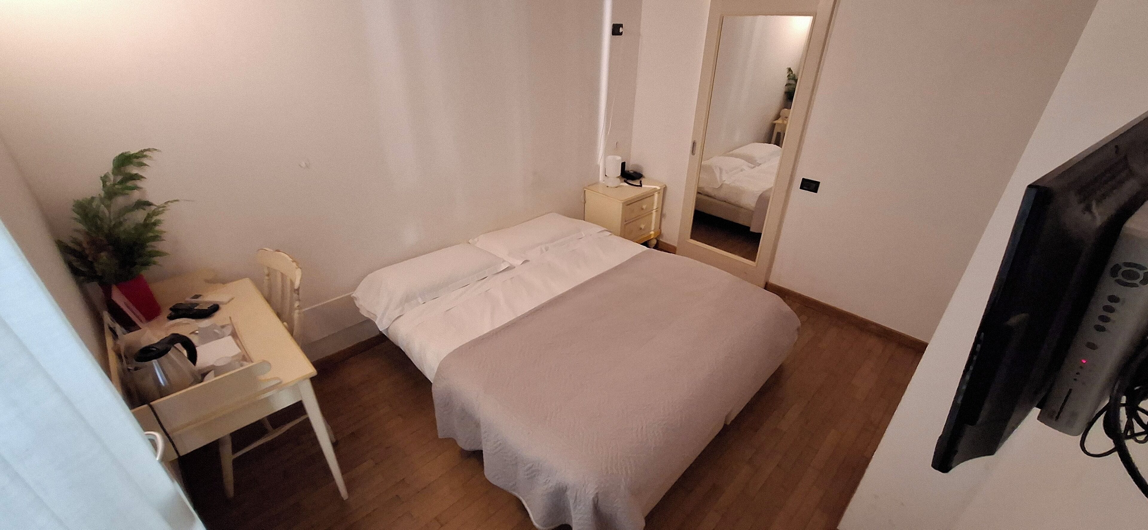 A small, neatly arranged bedroom with a single bed, a bedside table, a chair, a desk, and a television.