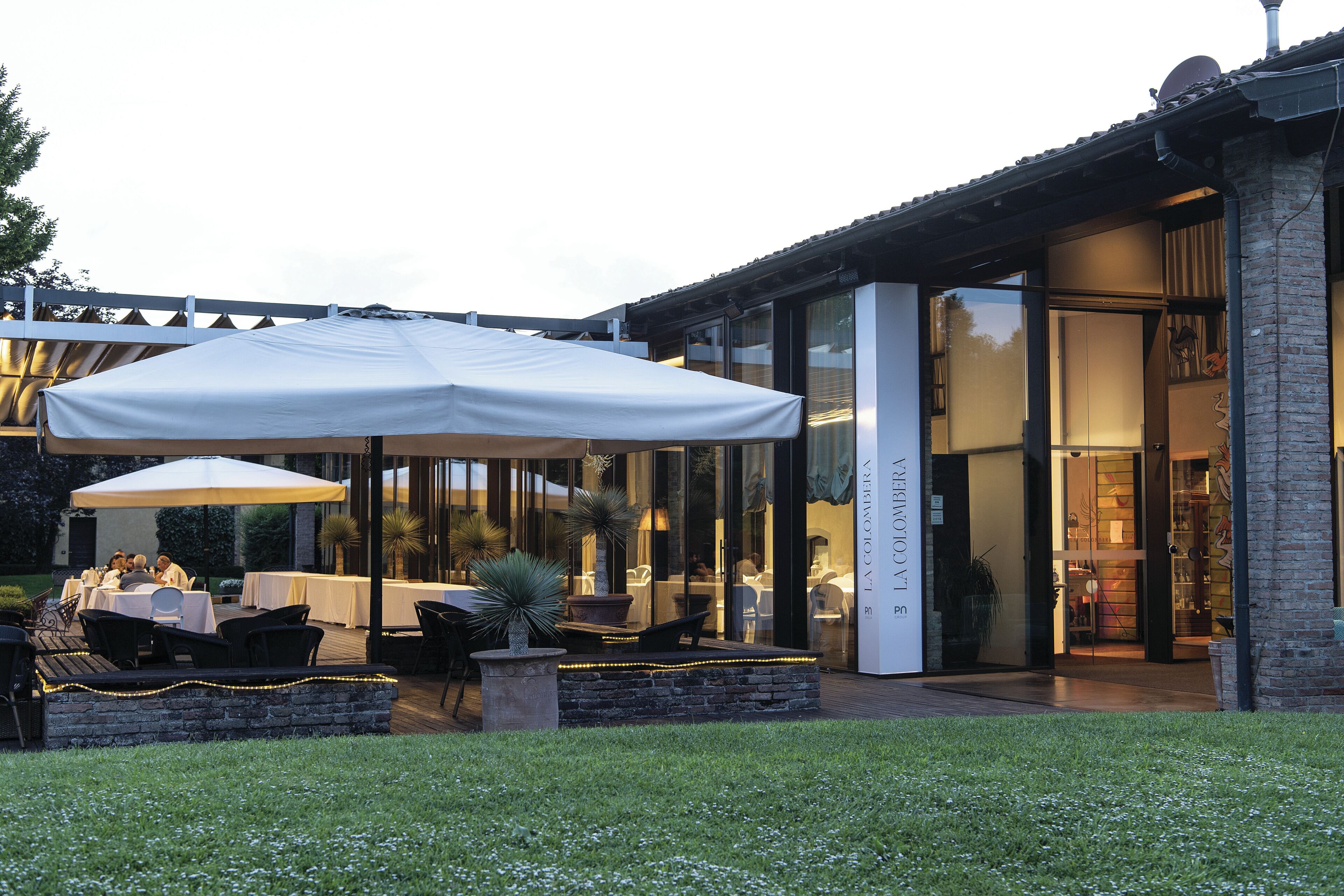 An outdoor dining area with tables and chairs, a large umbrella, and a building entrance with glass doors.