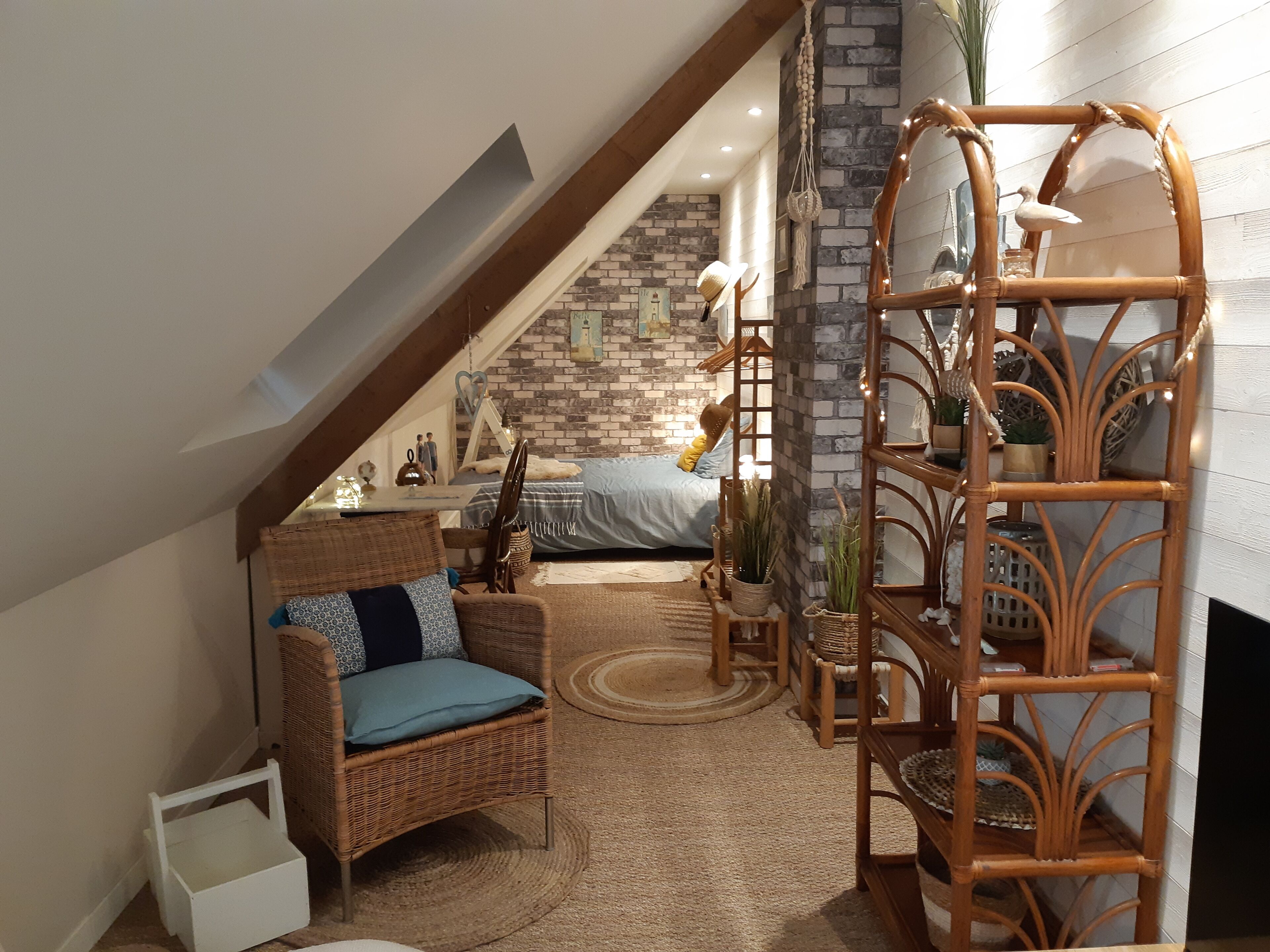 A cozy loft bedroom with a sloped ceiling, a brick accent wall, a bed with a blue bedspread, a wicker chair, a wooden shelf with plants, and a small white table with decorative items.