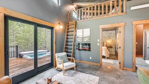 A spacious interior with a large sliding glass door leading to an outdoor hot tub area, a wooden staircase, and a cozy seating area with a view of the outdoors. - Secluded Modern Cabin- Near Flathead Lake (Polson)