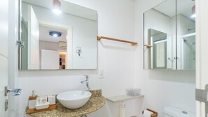 Une salle de bain moderne avec un lavabo blanc, un comptoir en granit et un miroir au-dessus du lavabo. On aperçoit un porte-serviettes au-dessus du miroir et des toilettes à l’arrière-plan.