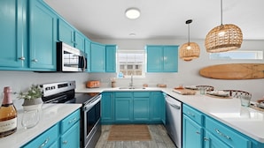 A modern kitchen with teal cabinets, stainless steel appliances, a wooden surfboard decoration, and a wicker pendant light. - Spacious Beach Retreat, Pool, Hot Tub, Sleeps 14 (Panama City Beach)