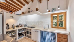A kitchen with wooden beams, a window with a view, built-in appliances, and a checkered towel. - CAS FORNER - Finca amb piscina a la Colònia (Colonia de Sant Pere)
