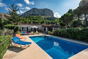 Une piscine entourée de chaises longues, un bâtiment avec un toit en tuiles et une montagne en arrière-plan.