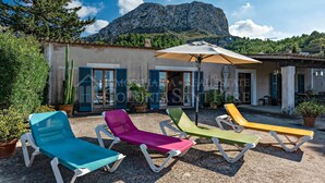 A patio with colorful sun loungers and a large umbrella, a building with a mountain backdrop, and a cactus plant. - CAS FORNER - Finca amb piscina a la Colònia (Colonia de Sant Pere)