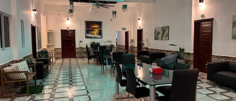 A hotel lobby with a tiled floor, seating areas, a glass table, ceiling fans, and hanging plants.