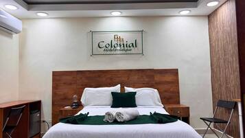 A hotel room with a wooden headboard, a bed with white linens and green pillows, a desk with a chair, and a nightstand with a lamp.