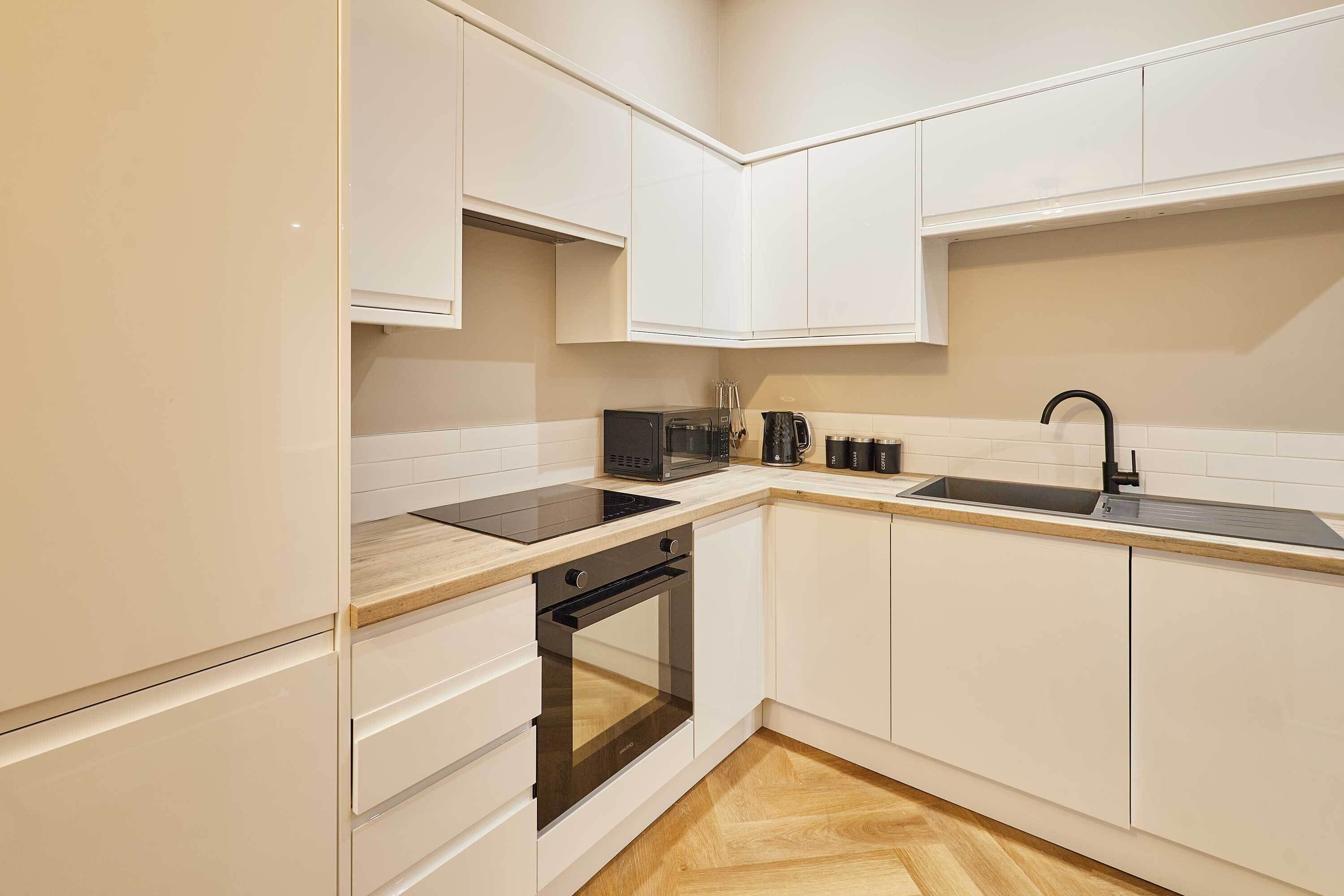 Une cuisine moderne avec des meubles blancs, une plaque de cuisson à induction noire, un four encastré, un four à micro-ondes, une machine à café et un évier.