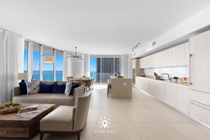 A modern living room with a sofa, coffee table, and dining area. A kitchen with white cabinets and a view of the city is visible. - Oceanline 3BR  Corner Balcony & Calm Seas (Hollywood)