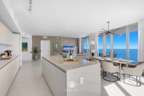 A modern hotel room with a kitchen area, dining space, and a large window with a view of the sea. - Oceanline 3BR  Corner Balcony & Calm Seas (Hollywood)