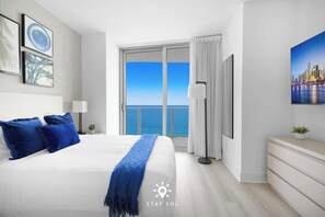 A hotel room with a large bed, a view of the sea, and a cityscape painting on the wall. - Oceanline 3BR  Corner Balcony & Calm Seas (Hollywood)