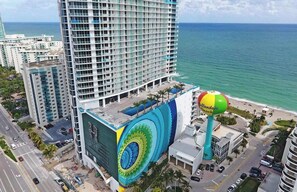 Un grand hôtel, avec une fresque murale imposante sur un de ses côtés, situé près d’une plage où se trouve une sculpture colorée en forme de ballon de plage.