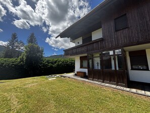 Une maison en bois à deux étages, dotée d’un balcon, d’une cour verdoyante et surplombant un paysage montagneux.