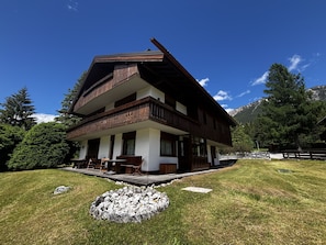 A two-story wooden chalet with a balcony, a stone fire pit, and a view of mountains.
