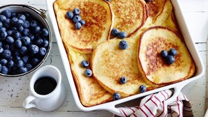 Un plat de cuisson rectangulaire blanc contient des crêpes dorées, garnies de myrtilles. À côté, on aperçoit un petit bol de myrtilles et un pichet blanc rempli de café, le tout posé sur une surface en bois.