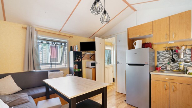 A compact kitchen with wooden cabinets, a refrigerator, and a dining area with a table and sofa. - Mobile Home "Mh 44 La Tremblade" with Shared Pool, Private Terrace, and Air Conditioning (La Tremblade)