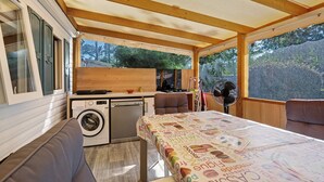 A compact kitchen with a washing machine, a dining table with a colorful tablecloth, and a fan. - Mobile Home "Mh 44 La Tremblade" with Shared Pool, Private Terrace, and Air Conditioning (La Tremblade)