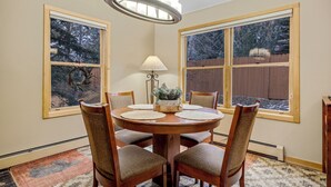 A dining room with a round wooden table, six chairs, a chandelier, and two windows with a view of a wooden fence and trees. - Cozy Keystone Townhome - walkable to both base areas! (Keystone)