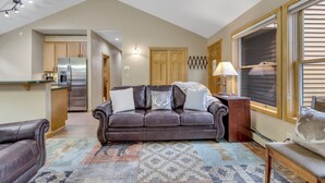 A spacious living area with a brown leather sofa, a patterned rug, and a wooden coffee table. A kitchen with wooden cabinets and a stainless steel refrigerator is visible in the background. - Cozy Keystone Townhome - walkable to both base areas! (Keystone)