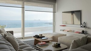 Un salon moderne doté d’une grande porte-fenêtre coulissante donnant sur un balcon avec vue sur la mer. On y trouve un canapé, une table basse et un téléviseur fixé au mur.