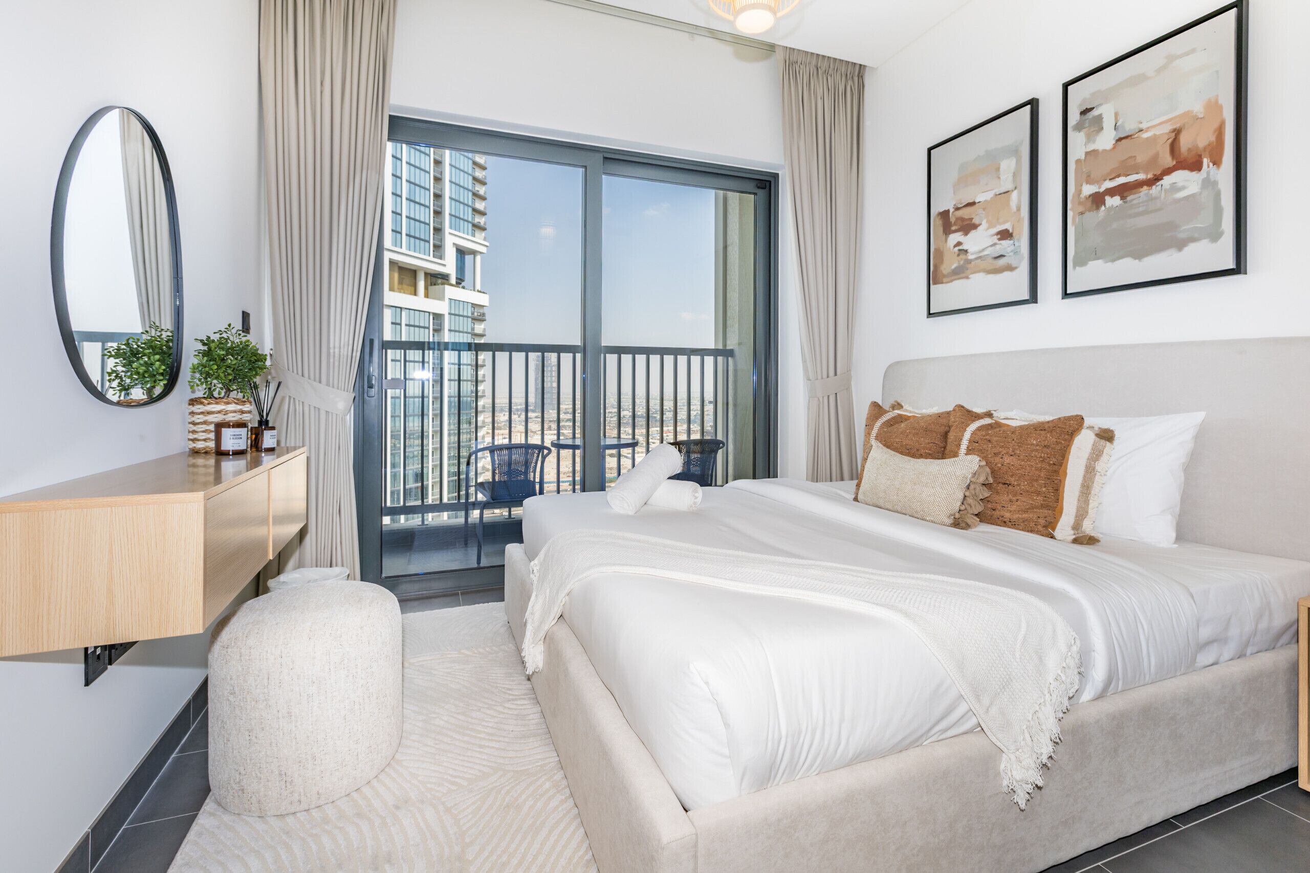 A modern bedroom with a large bed, a wooden bedside table, a round ottoman, and a view of a building through the window.