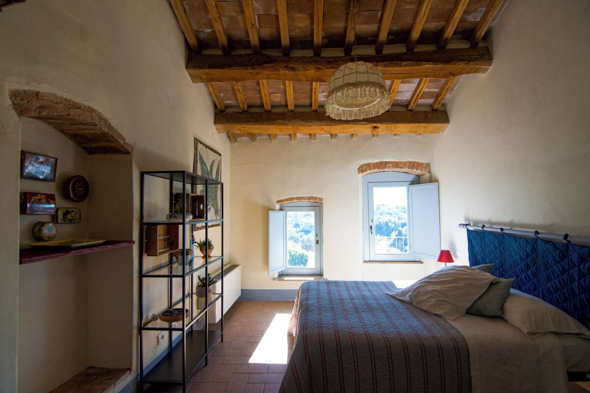 Ein Schlafzimmer mit einem Bett, einem Fenster, einer Ablage mit Dekoration und einer Decke mit sichtbaren Balken.