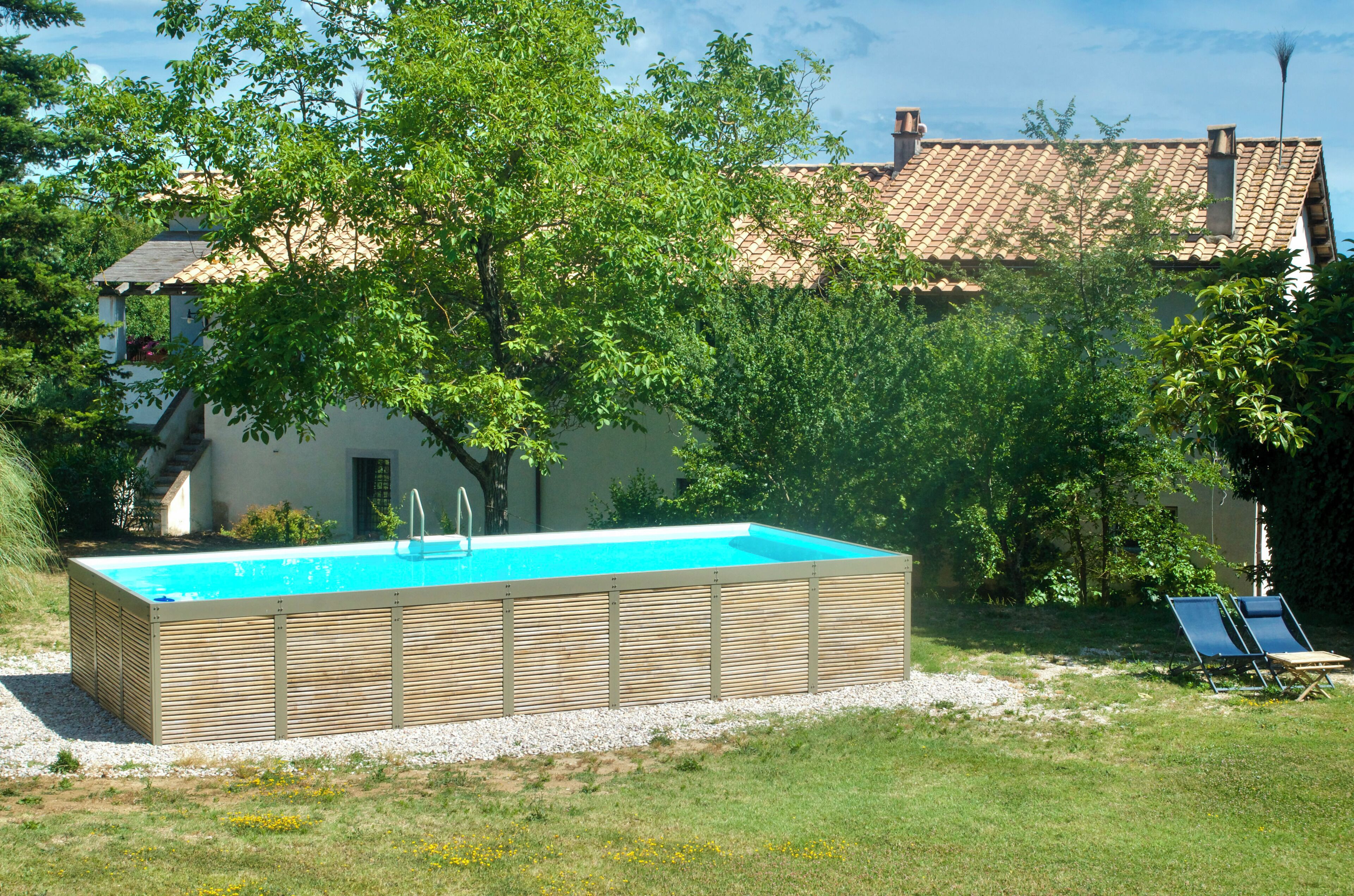 Una piscina rectangular con cubierta de madera, rodeada de césped y árboles, con un edificio de techo de tejas al fondo.