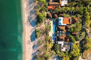 Vista aérea de uma propriedade costeira com piscina, vários edifícios e uma praia.