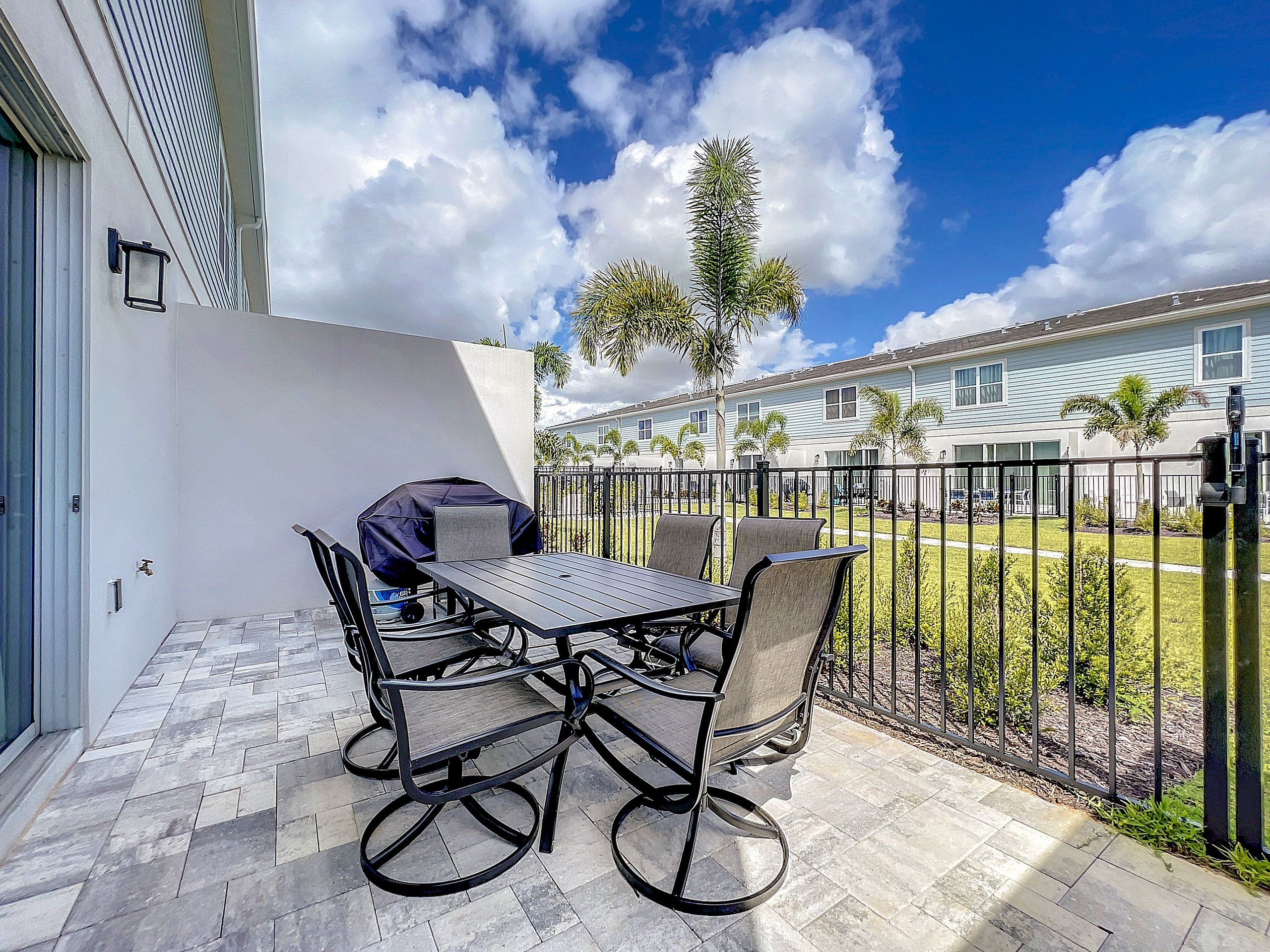 A patio with a table and chairs, a covered grill, and a view of a residential area with houses and palm trees.