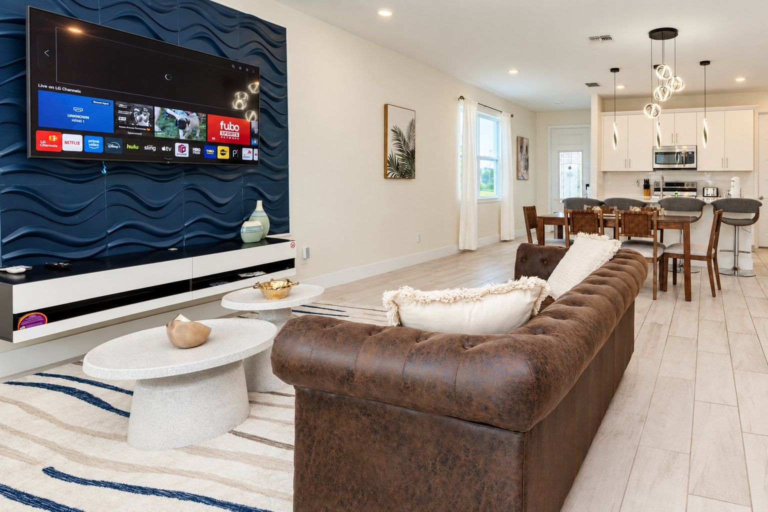 A modern living room with a flat-screen TV, a brown leather sofa, a dining area with a table and chairs, and a kitchen in the background.