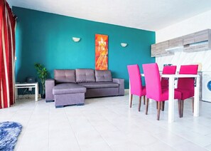Una sala de estar moderna con una pared de acento color verde azulado, un sofá gris, una mesa de comedor con sillas rosadas y una lavadora al fondo.