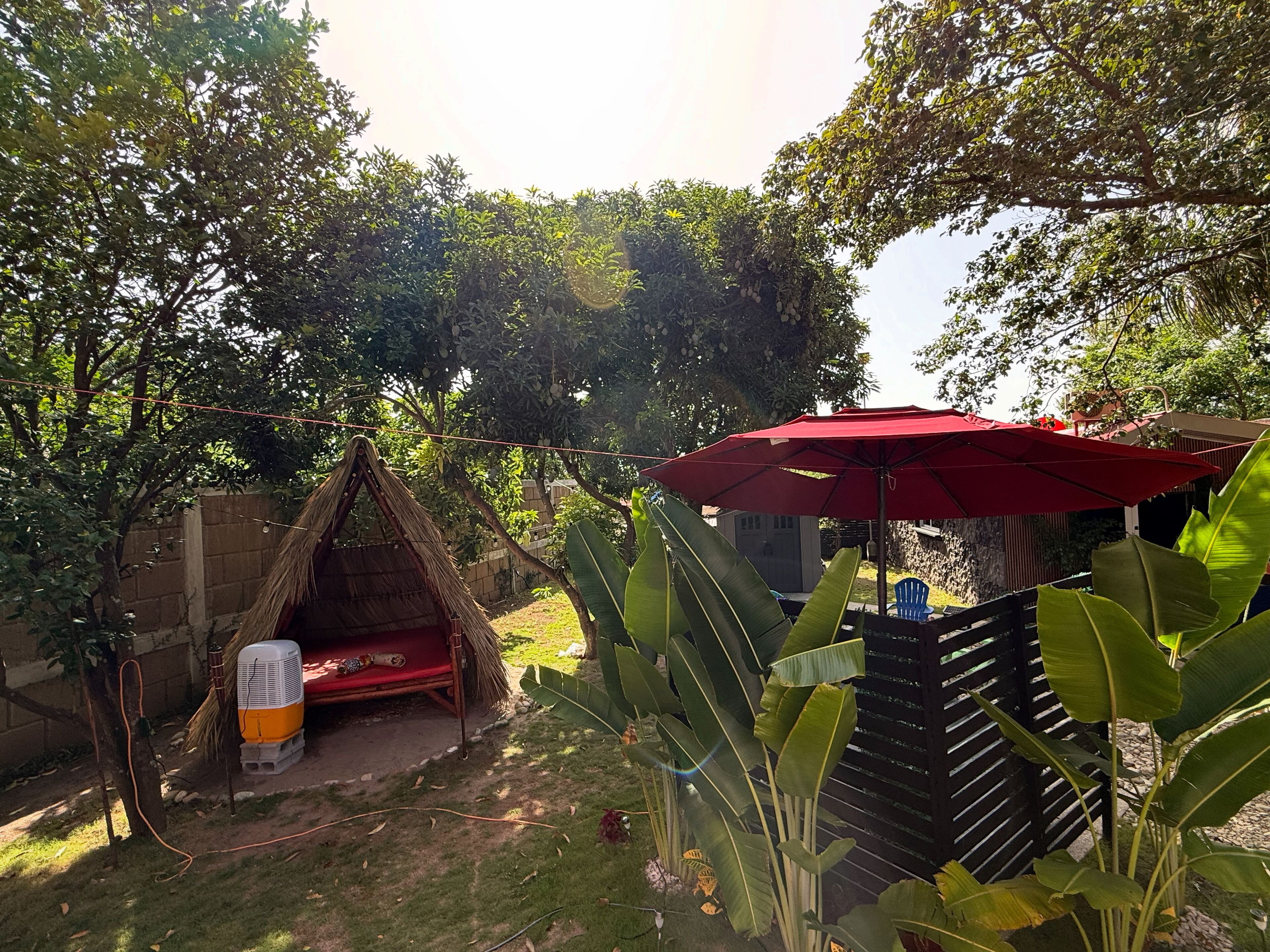 A thatched-roof hut with a fan, a red umbrella, and lush greenery.
