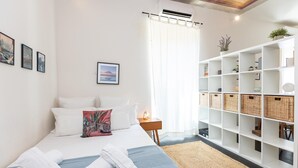 Una camera da letto ordinata con un letto, cuscini, un comodino, uno scaffale con cesti e una finestra con tende.