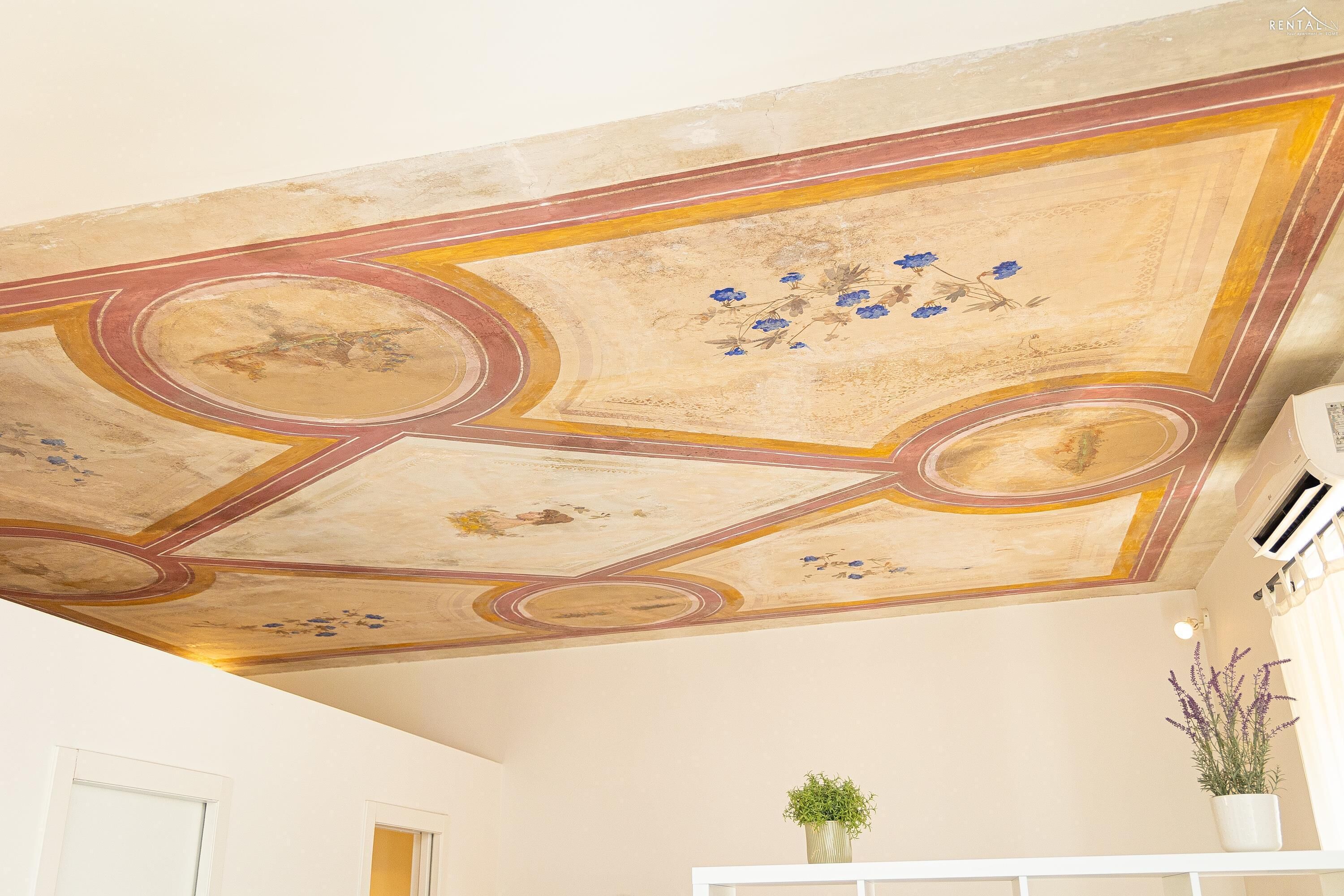 A room with a ceiling featuring a decorative fresco with floral patterns and geometric designs.