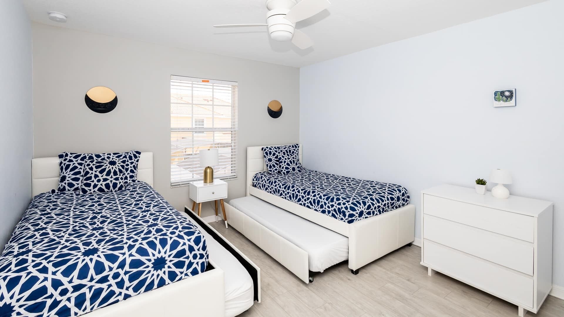 Une chambre moderne avec deux lits, une commode blanche, un banc et un ventilateur de plafond.